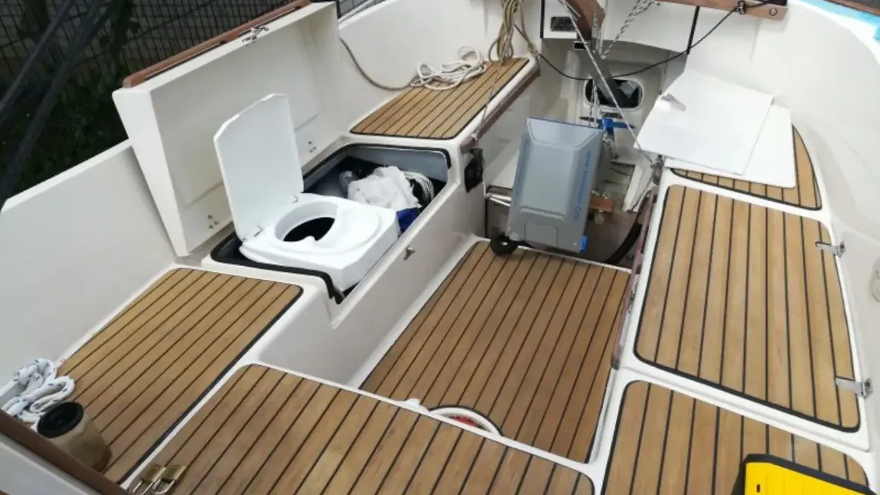 Ein weißes **Segelboot Toilette** mit offenem Deckel, daneben ein grauer Tank. Holzdecks und weiße Wände.