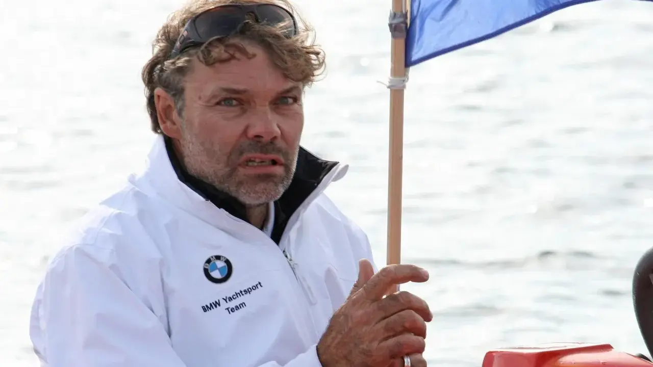 Markus Wieser, mit zerzaustem Haar und Sonnenbrille auf dem Kopf, trägt eine weiße BMW Yachtsport Team Jacke. Er blickt konzentriert auf das Wasser.