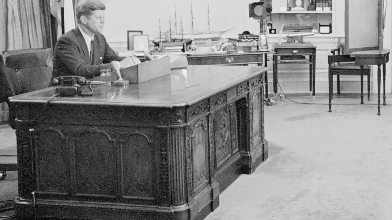 John F. Kennedy am Schreibtisch im Oval Office. Hinter ihm ein Modellschiff, das an die HMS Resolute erinnert, die dem Schreibtisch seinen Namen gab.