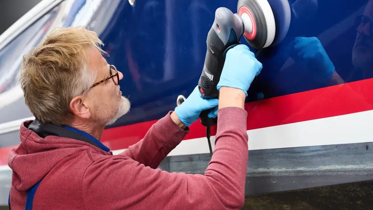 Mann mit Brille poliert mit Maschine den blauen Rumpf eines Bootes.