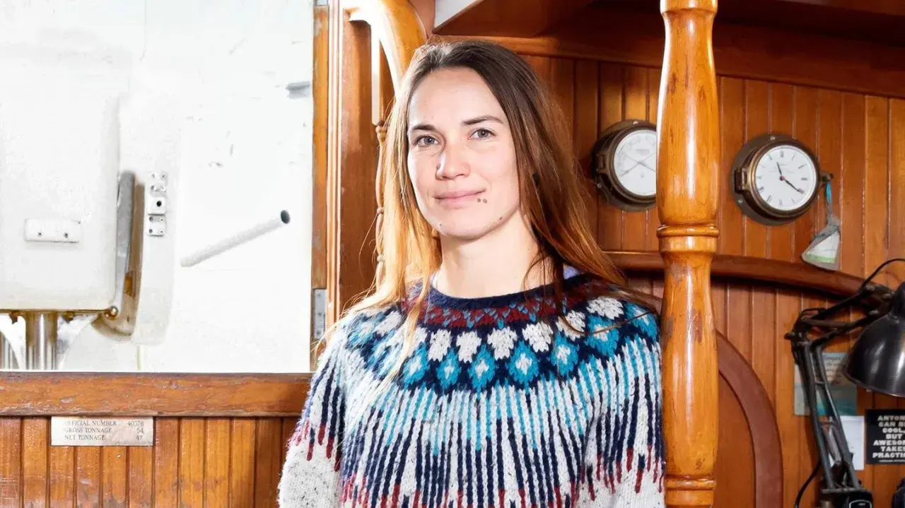 Laura Dekker heute, lächelnd in einem bunten Strickpullover, an Bord eines Schiffes mit Holzakzenten und maritimen Uhren.