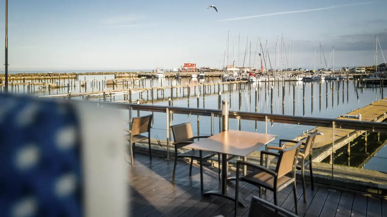 Ein Tisch mit Stühlen auf einer Holzterrasse mit Blick auf einen Yachthafen. Segelboote liegen im Wasser, die Sonne wirft lange Schatten. Ein Möwe fliegt über den Hafen. Damp 2000.
