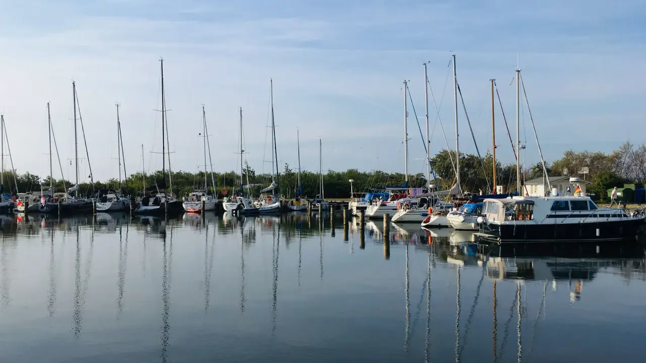 Segelboote und Motorboote liegen friedlich im Hafen Neuendorf, ihre Spiegelbilder tanzen auf dem ruhigen Wasser.