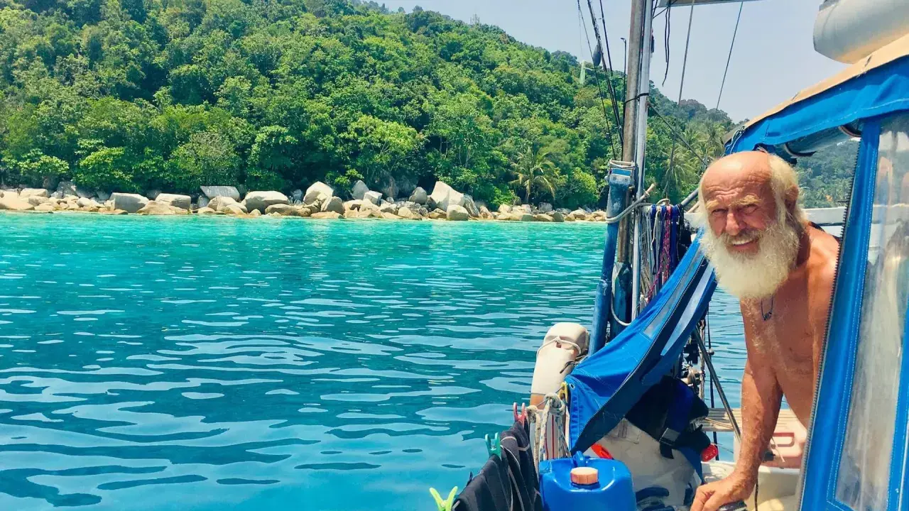 Gangerl Clemens genießt die Fahrt auf seinem Segelboot vor einer üppig grünen Insel mit türkisfarbenem Wasser.