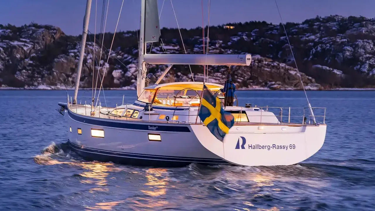 Eine Hallberg-Rassy 69 gleitet bei Dämmerung über das Wasser, die schwedische Flagge weht im Wind.