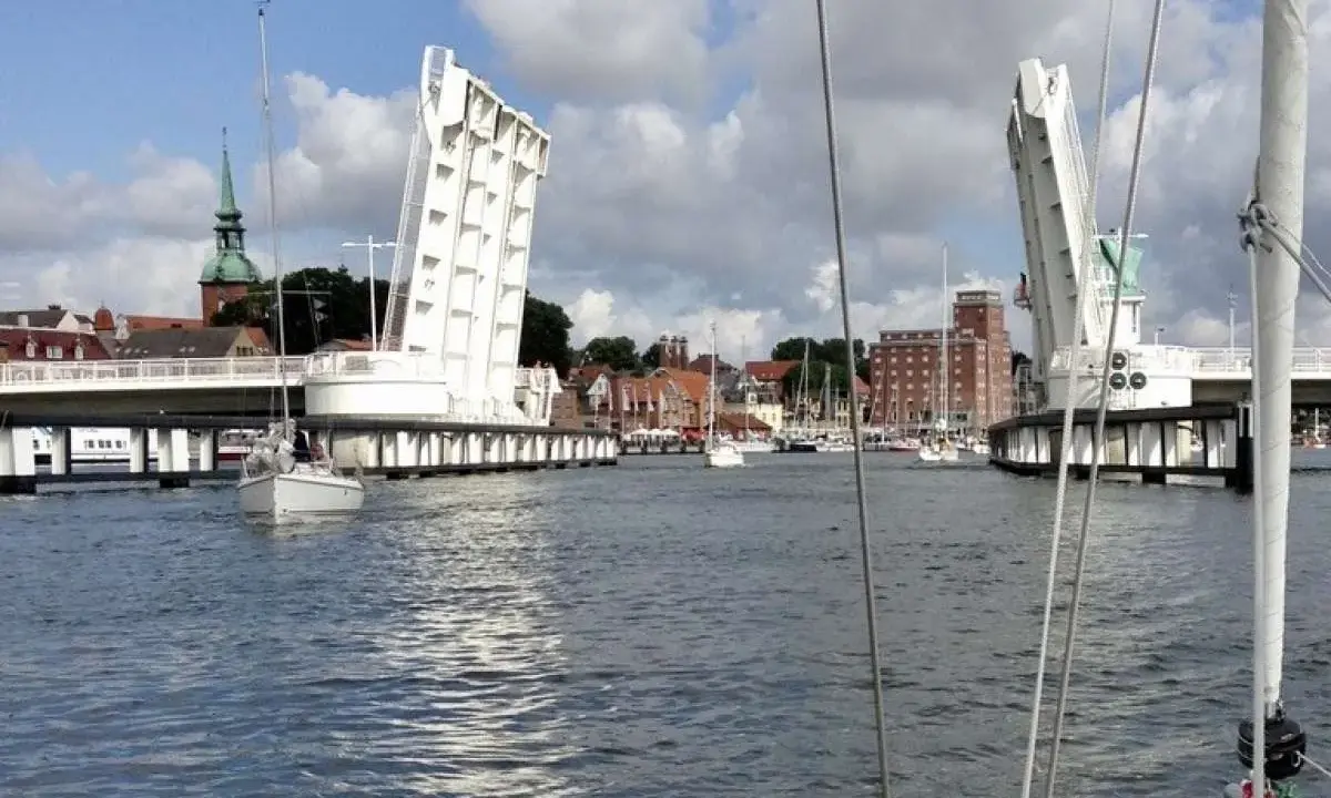 Eine Klappbrücke öffnet sich für Segelboote in Kappeln an der Schlei. Im Hintergrund die Stadt mit Kirchturm und Backsteingebäuden.