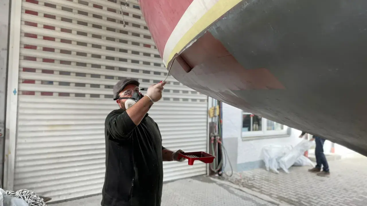Mann mit Maske und Schutzbrille streicht mit einer Rolle den Rumpf eines Bootes.
