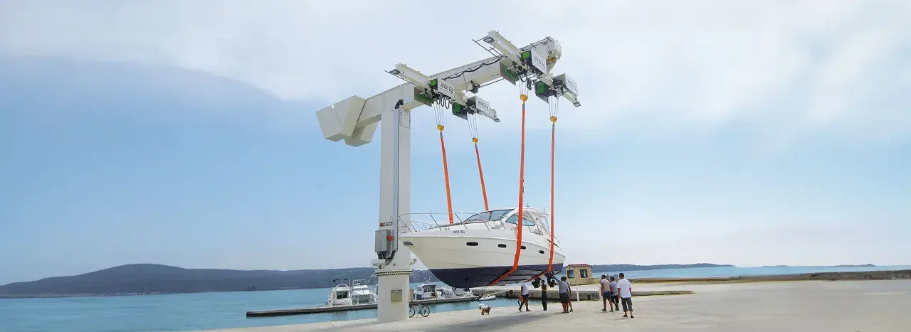Ein großer Kran hebt ein weißes Boot aus dem Wasser. Mehrere Personen beobachten den Vorgang.