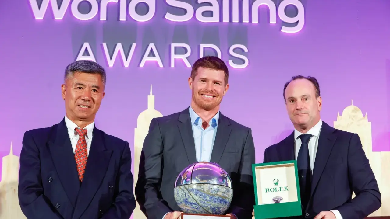Tom Slingsby mit Auszeichnung bei den World Sailing Awards. Er hält eine Trophäe, daneben zwei Männer mit Geschenken.