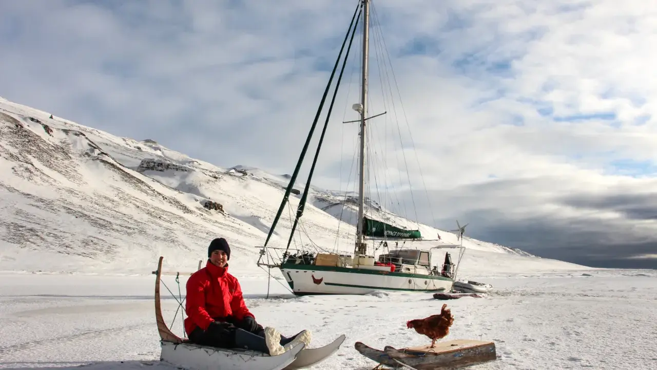 Guirec Soudée sitzt in einem Schlitten vor seinem Segelboot in einer verschneiten Landschaft. Eine Henne steht auf einem Holzbrett daneben.