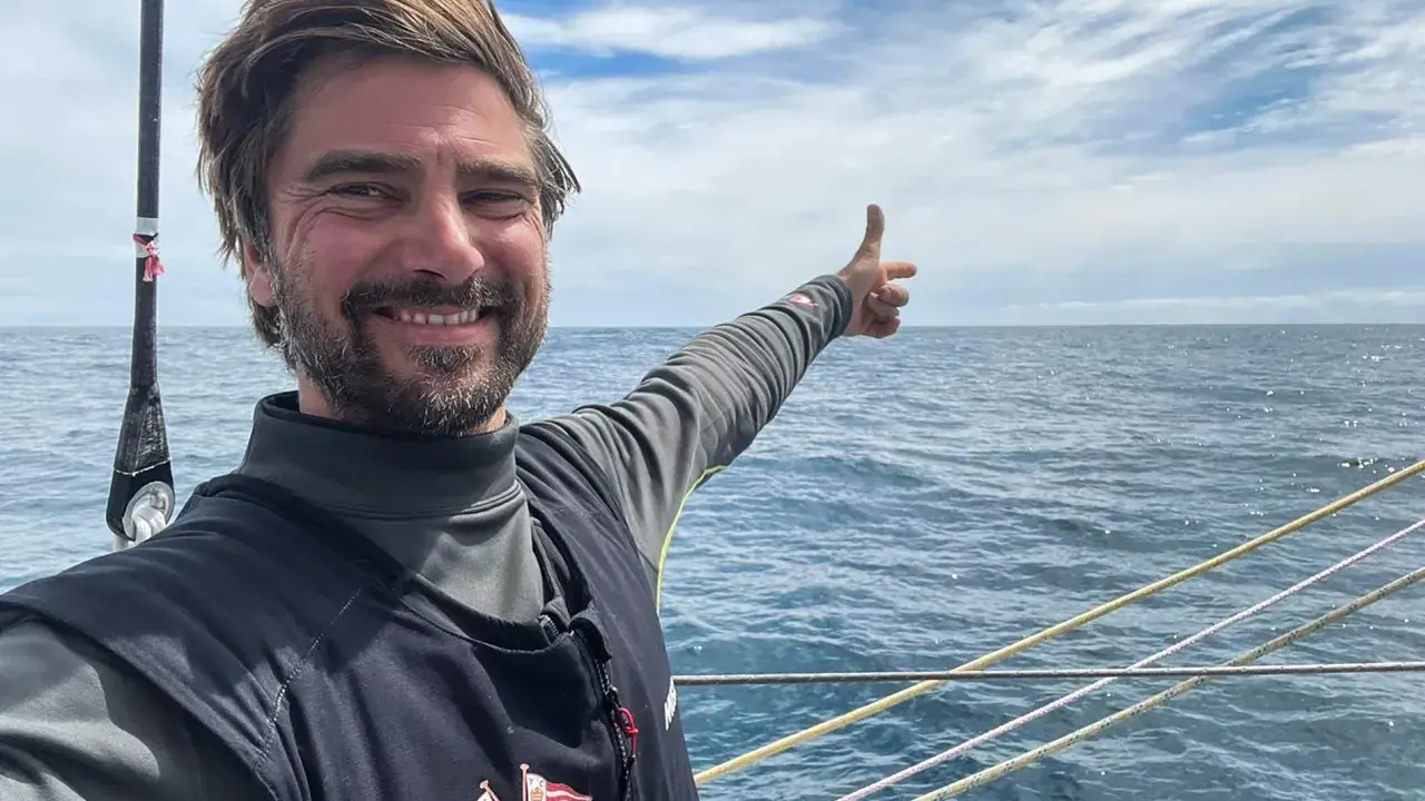 Boris Herrmann zeigt auf das weite Meer, ein Lächeln im Gesicht. Er ist auf einem Segelboot unterwegs, die Wellen glitzern.