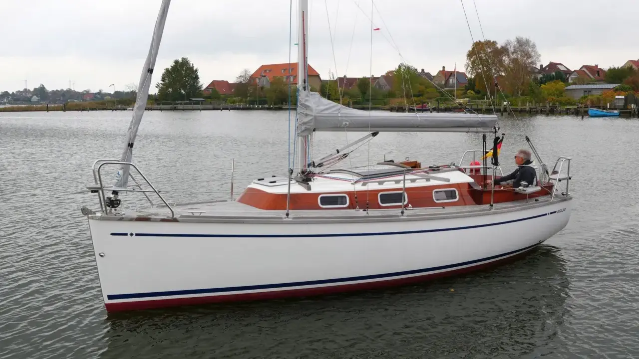 Ein Mann steuert die Segelyacht BIGA 242 auf ruhigem Wasser, im Hintergrund Häuser und Bäume.