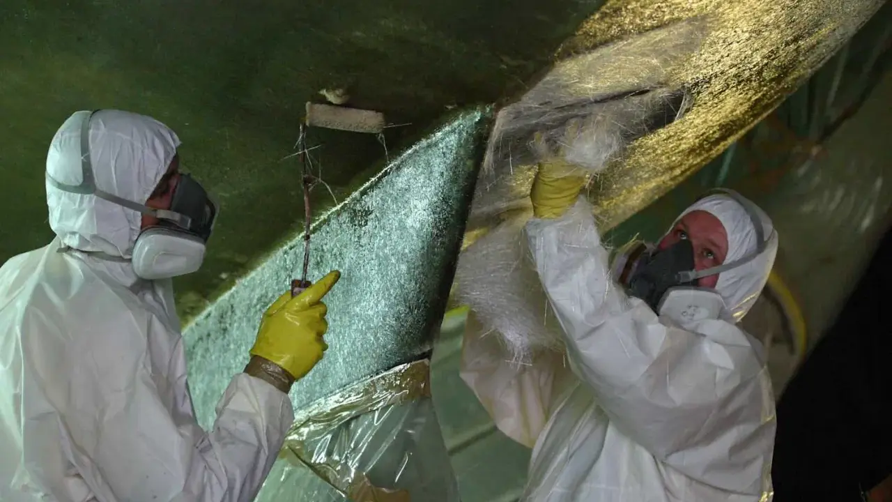 Zwei Personen in Schutzkleidung reparieren den Rumpf eines Bootes. Sie tragen Glasfasermatten auf, um Osmose-Schäden zu beheben.