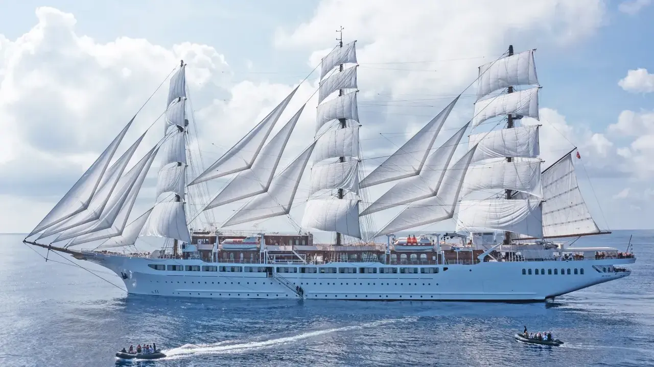 Ein majestätisches Segelschiff mit vollen weißen Segeln, das wie ein **sea cloud spirit** über das blaue Meer gleitet. Kleine Beiboote sind in der Nähe.