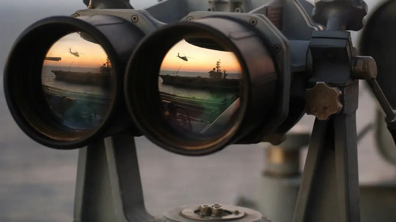 Fernglas-Werte: Ein Flugzeugträger und ein Hubschrauber spiegeln sich im Okular eines Fernglases bei Sonnenuntergang.