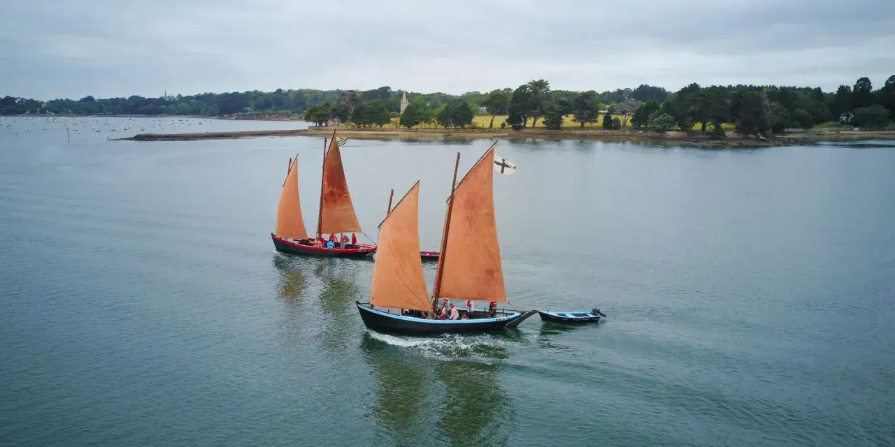 Drei traditionelle Segelboote mit orangefarbenen Segeln auf dem Wasser vor der Küste der Bretagne.