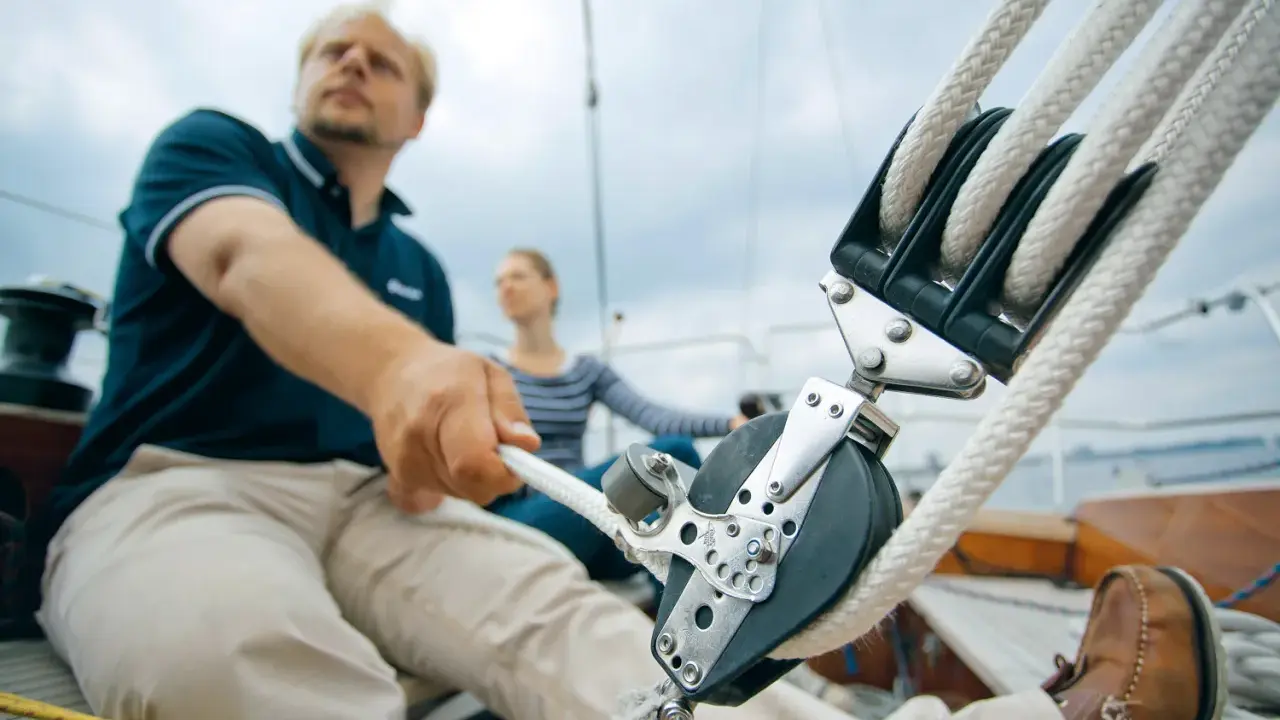 Mann zieht mit kräftiger Hand das Tauwerk am Segel. Ein Mann und eine Frau auf einem Segelboot, die das Tauwerk am Segel bedienen.