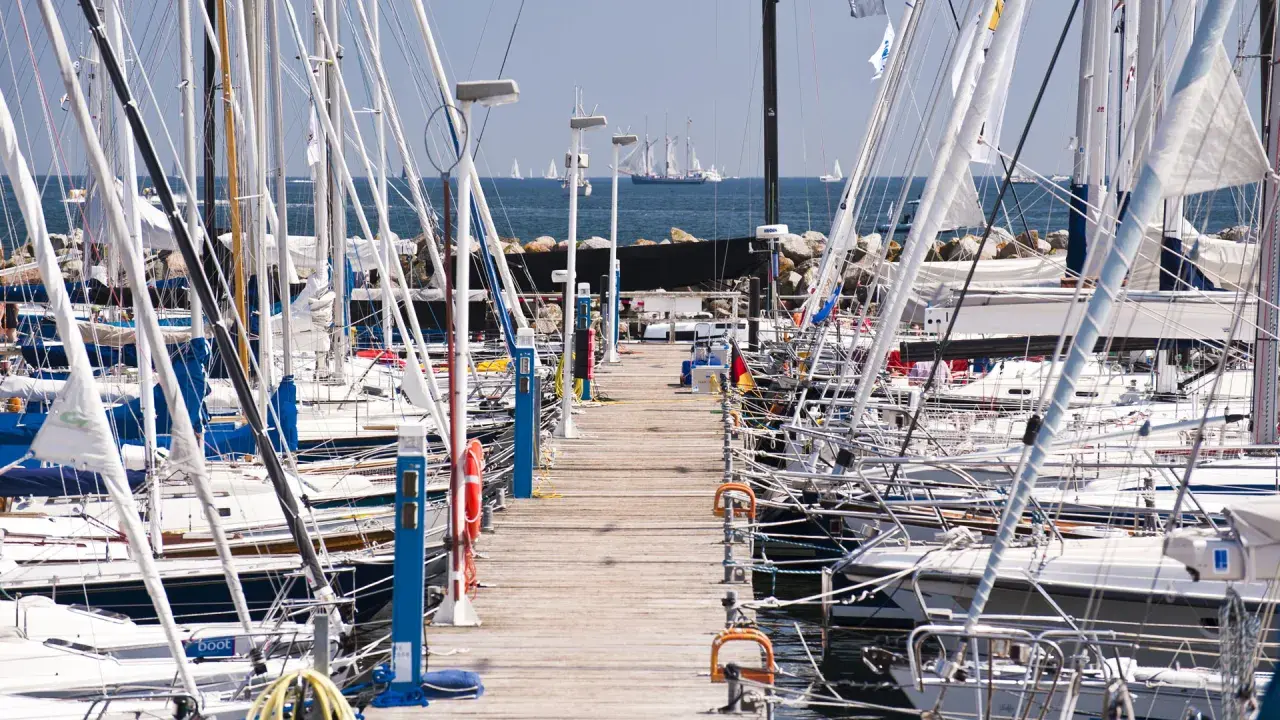 Viele Segelboote liegen im **Flensburg Hafen** an einem Holzsteg. Im Hintergrund segeln große Schiffe auf dem Meer.