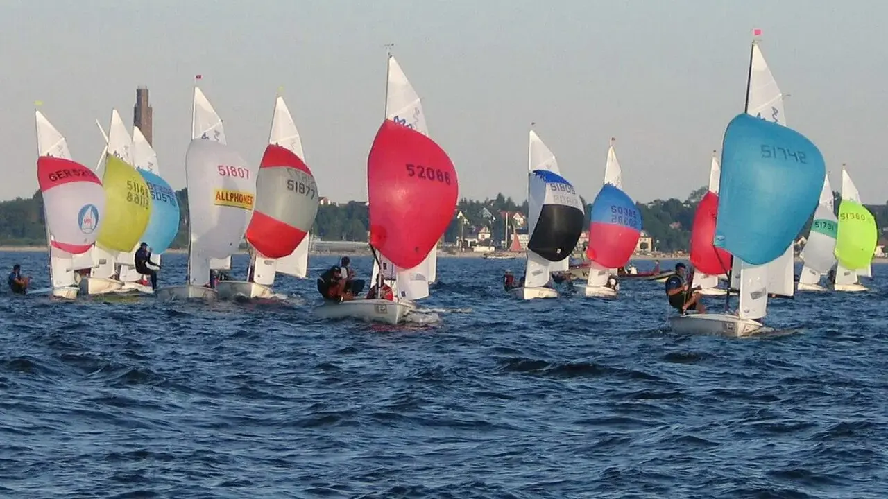 Bunte Segel dominieren das Bild einer spannenden Regatta. Mehrere kleine Boote mit Seglern sind auf dem Wasser unterwegs.