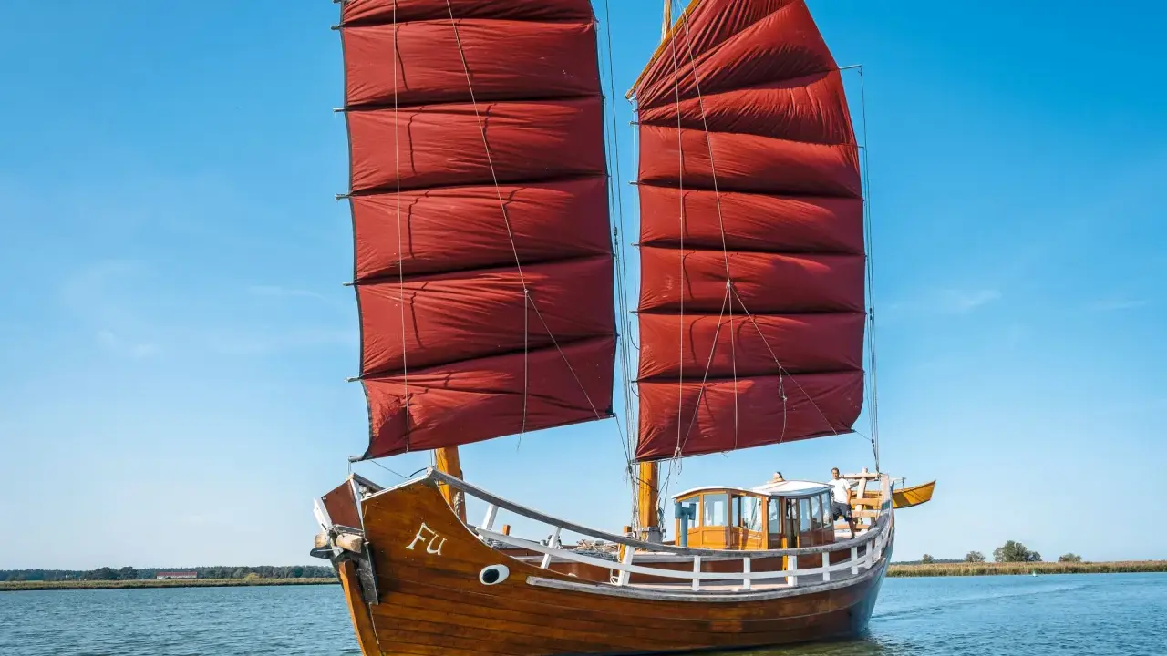 Ein hölzernes Dschunke Schiff mit roten Segeln gleitet auf ruhigem Wasser unter blauem Himmel.