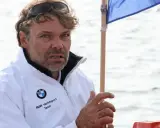 Markus Wieser, mit zerzaustem Haar und Sonnenbrille auf dem Kopf, trägt eine weiße BMW Yachtsport Team Jacke. Er blickt konzentriert auf das Wasser.