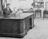 John F. Kennedy am Schreibtisch im Oval Office. Hinter ihm ein Modellschiff, das an die HMS Resolute erinnert, die dem Schreibtisch seinen Namen gab.