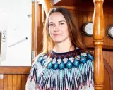 Laura Dekker heute, lächelnd in einem bunten Strickpullover, an Bord eines Schiffes mit Holzakzenten und maritimen Uhren.