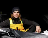 Clarisse Crémer, Seglerin im L'Occitane Sailing Team, sitzt in dunkler Kleidung und gelber Latzhose inmitten von Segeln.