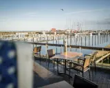 Ein Tisch mit Stühlen auf einer Holzterrasse mit Blick auf einen Yachthafen. Segelboote liegen im Wasser, die Sonne wirft lange Schatten. Ein Möwe fliegt über den Hafen. Damp 2000.