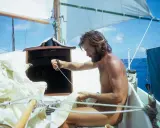 Wilfried Erdmann, mit Bart und langen Haaren, arbeitet an Bord einer Segelyacht. Er zieht an einer Leine, während die Segel im Wind wehen.