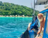 Gangerl Clemens genießt die Fahrt auf seinem Segelboot vor einer üppig grünen Insel mit türkisfarbenem Wasser.