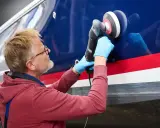 Mann poliert mit Maschine den blauen Bootsrumpf. Das Gelcoat erstrahlt nach dem Polieren wieder in neuem Glanz.