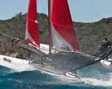 Zwei Segler auf einem Katamaran mit roten Segeln, der über die Ostsee rast. Gischt spritzt auf, während sie mit voller Kraft segeln.