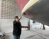 Mann mit Maske und Schutzbrille streicht mit einer Rolle den Rumpf eines Bootes.