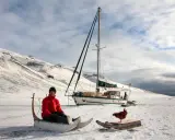 Guirec Soudée sitzt in einem Schlitten vor seinem Segelboot in einer verschneiten Landschaft. Eine Henne steht auf einem Holzbrett daneben.