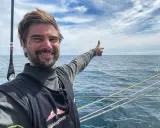Boris Herrmann zeigt auf das weite Meer, ein Lächeln im Gesicht. Er ist auf einem Segelboot unterwegs, die Wellen glitzern.
