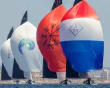Segelboote der j class rasen über das Meer. Bunte Spinnaker mit Logos und Symbolen, im Hintergrund eine Küstenstadt.