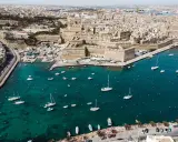 Segeln in Malta: Boote und Yachten im türkisfarbenen Wasser vor einer historischen Stadtkulisse mit Festungsmauern.