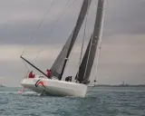 Ein IMOCA 60 segelt auf stürmischer See, die Crew in roten Jacken bereitet sich auf die nächste Welle vor.