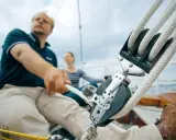Mann zieht mit kräftiger Hand das Tauwerk am Segel. Ein Mann und eine Frau auf einem Segelboot, die das Tauwerk am Segel bedienen.