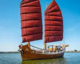 Ein hölzernes Dschunke Schiff mit roten Segeln gleitet auf ruhigem Wasser unter blauem Himmel.