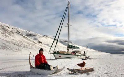 Guirec Soudée sitzt in einem Schlitten vor seinem Segelboot in einer verschneiten Landschaft. Eine Henne steht auf einem Holzbrett daneben.