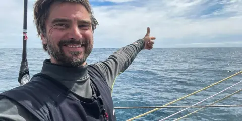 Boris Herrmann zeigt auf das weite Meer, ein Lächeln im Gesicht. Er ist auf einem Segelboot unterwegs, die Wellen glitzern.