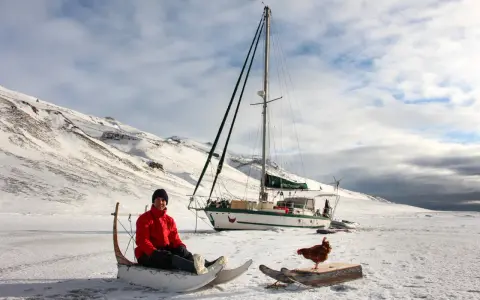 Guirec Soudée sitzt in einem Schlitten vor seinem Segelboot in einer verschneiten Landschaft. Eine Henne steht auf einem Holzbrett daneben.