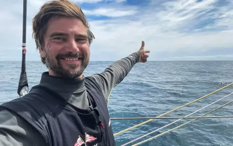 Boris Herrmann zeigt auf das weite Meer, ein Lächeln im Gesicht. Er ist auf einem Segelboot unterwegs, die Wellen glitzern.