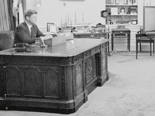 John F. Kennedy am Schreibtisch im Oval Office. Hinter ihm ein Modellschiff, das an die HMS Resolute erinnert, die dem Schreibtisch seinen Namen gab.
