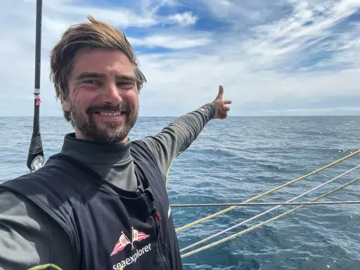 Boris Herrmann zeigt auf das weite Meer, ein Lächeln im Gesicht. Er ist auf einem Segelboot unterwegs, die Wellen glitzern.