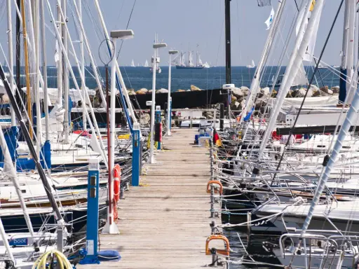 Viele Segelboote liegen im **Flensburg Hafen** an einem Holzsteg. Im Hintergrund segeln große Schiffe auf dem Meer.
