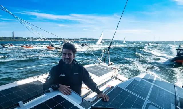 Boris Herrmann in Kiel, auf seinem Boot mit Solarpaneelen, umgeben von anderen Booten und Segelschiffen auf dem Meer.