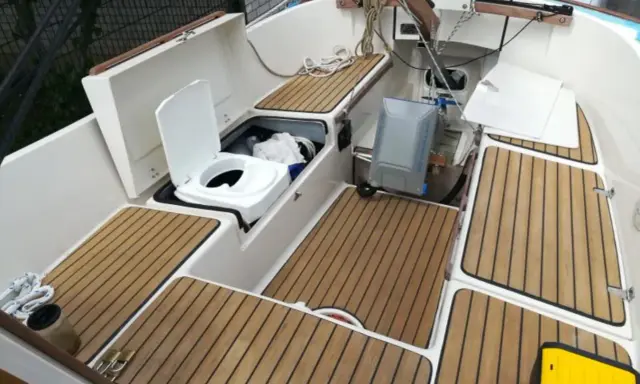 Ein weißes **Segelboot Toilette** mit offenem Deckel, daneben ein grauer Tank. Holzdecks und weiße Wände.